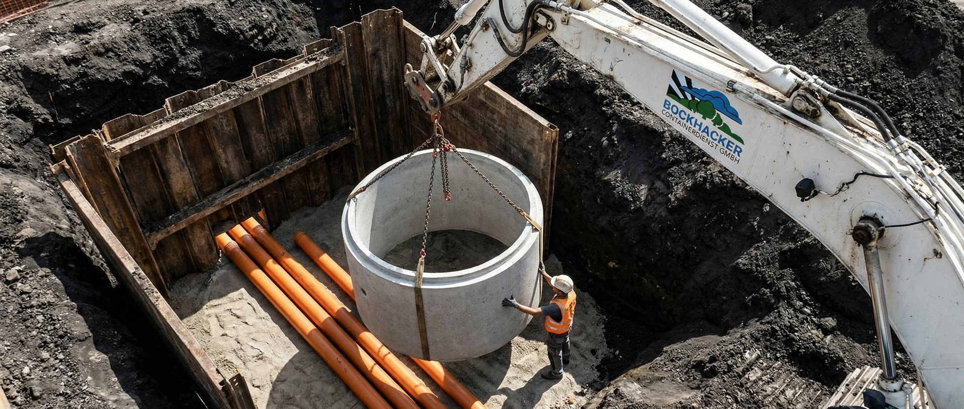 Professioneller Kanalbau und Erdarbeiten von Bockhacker Bagger setzt einen Betonschachtring für den Kanalbau in eine Baugrube, ein Arbeiter dirigiert ihn.